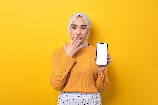 Bella ragazza asiatica stupita che indossa l'hijab che tiene il telefono cellulare con schermo bianco vuoto che copre la bocca con la mano isolata su sfondo giallo mockup copia spazio