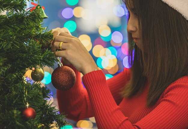 Bella ragazza asiatica sul maglione della santa che appende l'ornamento affascinante variopinto e la palla sull'albero di Natale per decorare la festa splendida di celebrazione nella notte beata. Aggiungi un po' di rumore per adattare l'immagine in stile vintage.