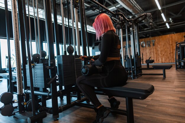 Bella ragazza atletica alla moda con i capelli rosa in abiti sportivi neri alla moda si siede vicino al simulatore e si prepara a fare esercizi in palestra