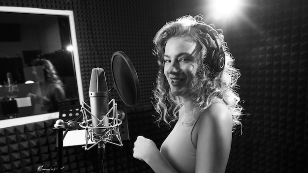 Bella ragazza bionda che canta emotivamente una canzone in studio di registrazione con microfono professionale e cuffie crea un nuovo album di brani artista vocale in bianco e nero girato in primo piano