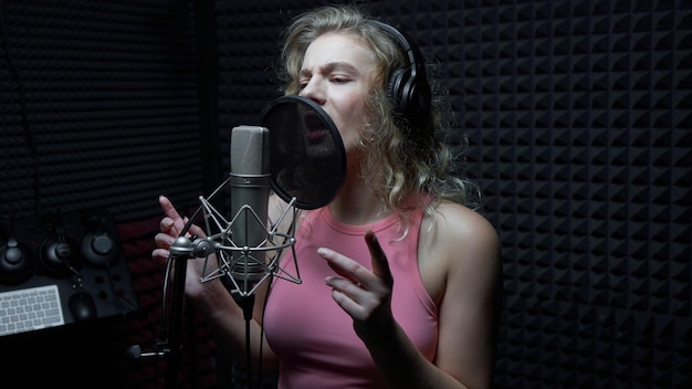 Bella ragazza bionda che canta emotivamente una canzone in studio di registrazione con microfono professionale e cuffie crea un nuovo album di tracce vocali artist closeup face