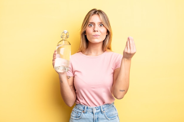 Bella ragazza bionda che fa un gesto capice o denaro, dicendoti di pagare. concetto di bottiglia d'acqua