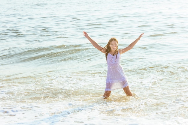 Bella ragazza bionda che indossa un abito bianco fluido in piedi alla caviglia nell'acqua dell'oceano