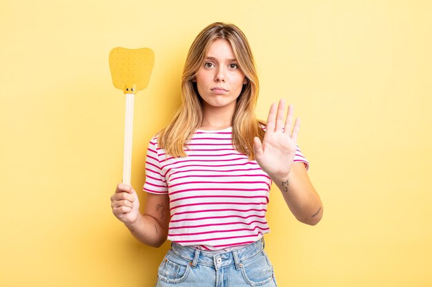 Bella ragazza bionda che sembra seria mostrando palmo aperto che fa gesto di arresto. uccidere mosche concetto