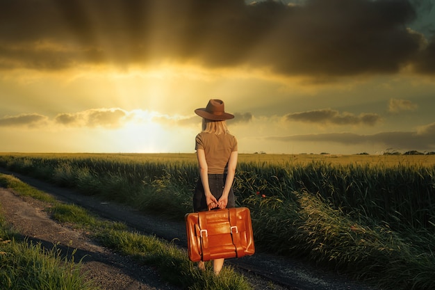 Bella ragazza bionda con la valigia in strada di campagna nell'ora del tramonto