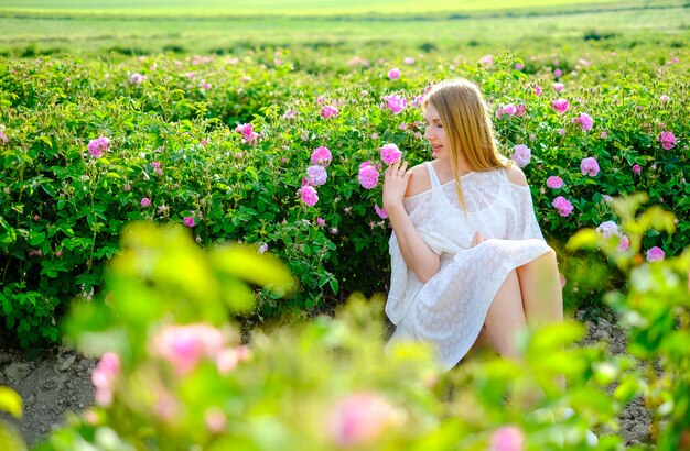 Bella ragazza bionda in abito bianco su una piantagione di rose in fiore