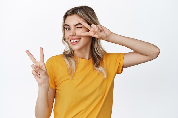 Bella ragazza bionda in posa con segni a V, che mostra gesto di pace e strizza l'occhio con un sorriso allegro, in piedi su bianco.