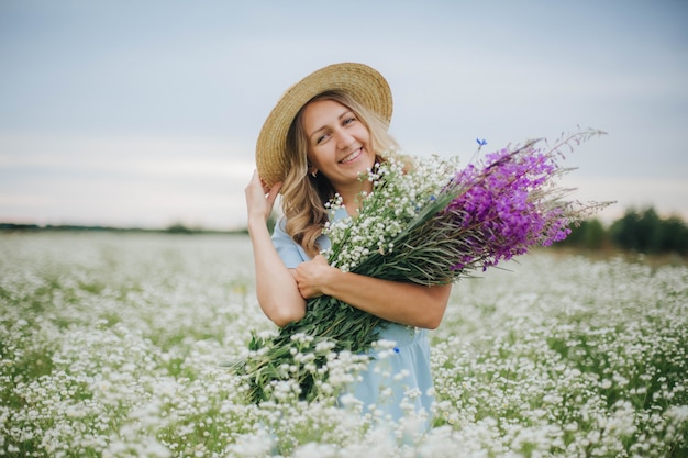 bella ragazza bionda in un campo di margherite. donna in un vestito blu in un campo di fiori bianchi. ragazza con un mazzo di margherite. foto tenera estiva nel villaggio. fiori selvatici. ragazza in un cappello di paglia