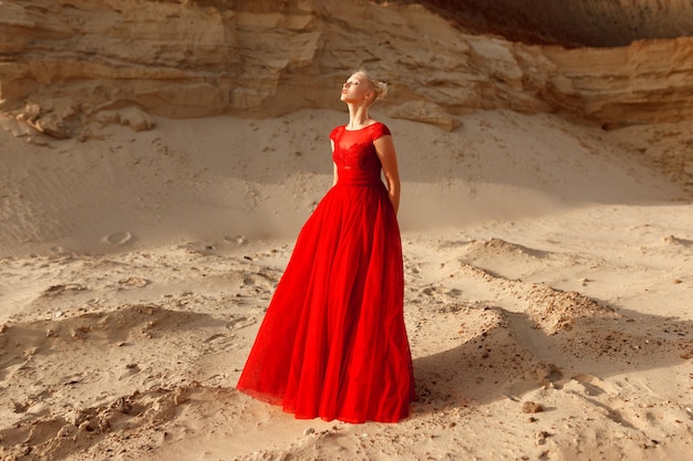 Bella ragazza bionda in un vestito rosso da principessa in posa nel deserto. Modello alla moda in posa in abito rosso al tramonto.