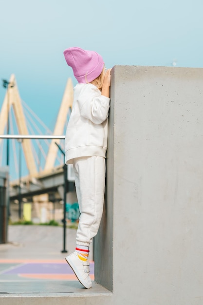 bella ragazza bionda in una tuta bianca e un cappello rosa si arrampica su una recinzione di cemento sullo sfondo di un paesaggio cittadino