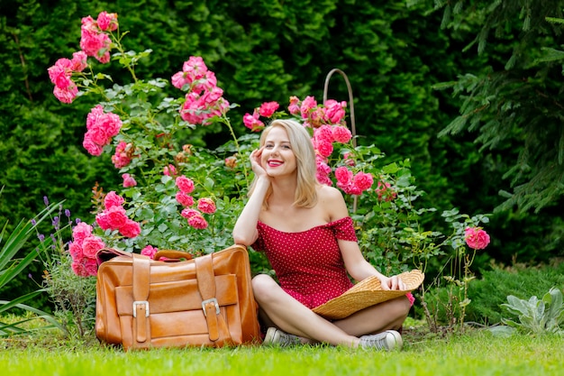 Bella ragazza bionda in vestito rosso con la valigia in un giardino