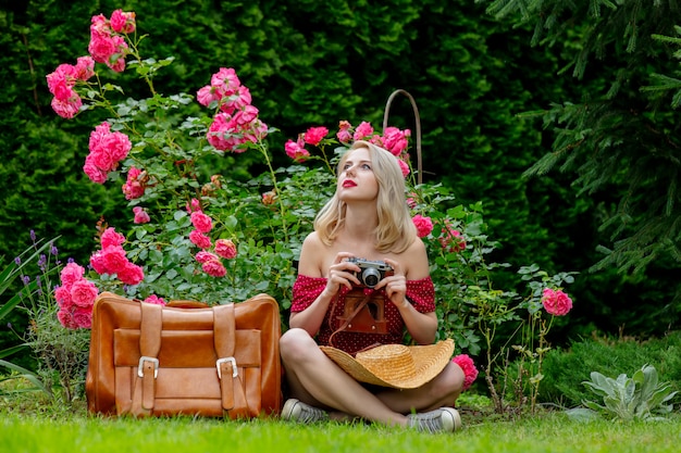 Bella ragazza bionda in vestito rosso con macchina fotografica d'epoca e valigia in un giardino