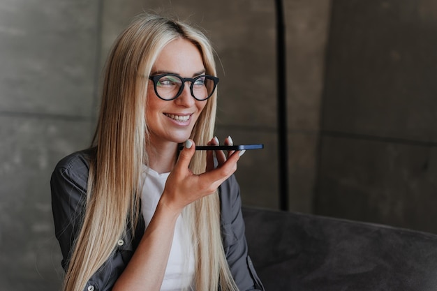 Bella ragazza bionda italiana con gli occhiali che registra un messaggio audio sorridente largamente vestita con un look casual