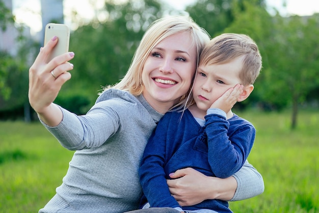 Bella ragazza bionda (madre) e figlio carino fanno selfie al telefono nel parco su uno sfondo di erba verde e alberi