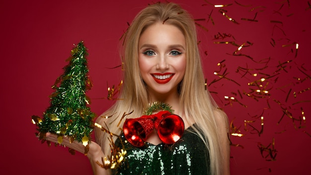 Bella ragazza bionda nell'immagine di un nuovo anno con campane di Natale al collo e albero verde. Fronte di bellezza con trucco festivo. Foto scattata in studio.