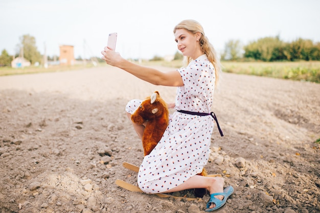 Bella ragazza bionda sembrante piacevole che fa la foto del selfie sul telefono. Giovane femmina graziosa felice nel retro cavallo alla moda del giocattolo da equitazione del vestito all'aperto.