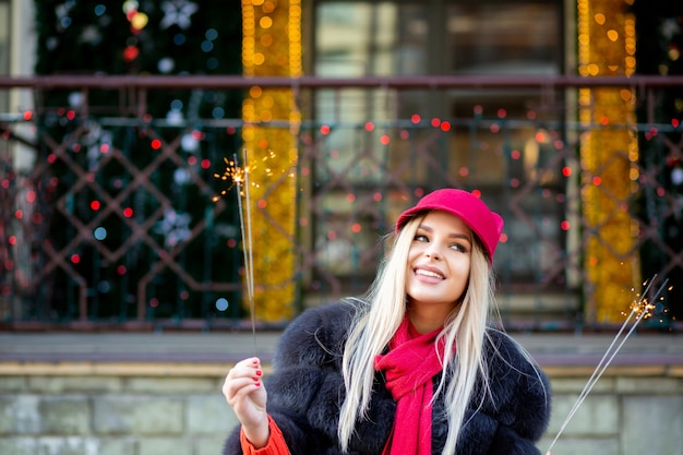 Bella ragazza bionda sorridente che indossa un cappello rosso alla moda che celebra il nuovo anno sullo sfondo del bokeh delle ghirlande. Spazio per il testo