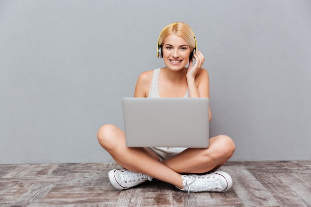 Bella ragazza bionda sorridente con il computer portatile e le cuffie isolate sul muro grigio