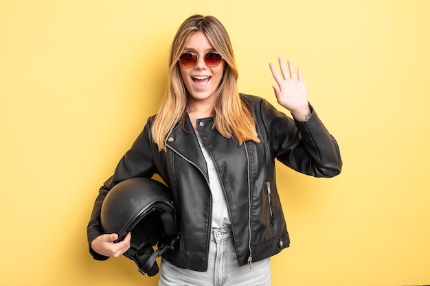 Bella ragazza bionda sorridente felicemente agitando la mano dandoti il benvenuto e salutandoti concetto di casco da moto