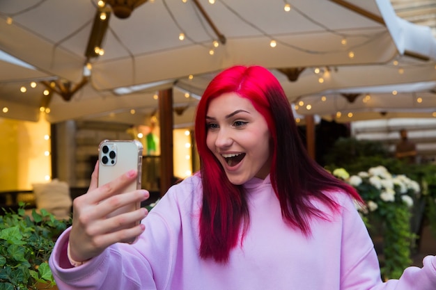 Bella ragazza blogger influencer dai capelli rossi al bar che parla facendo videochiamate utilizzando lo smartphone con i suoi abbonati nei social network.