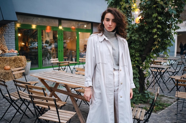 Bella ragazza bruna casual in trench che guarda con sicurezza a porte chiuse nella caffetteria sulla strada della città