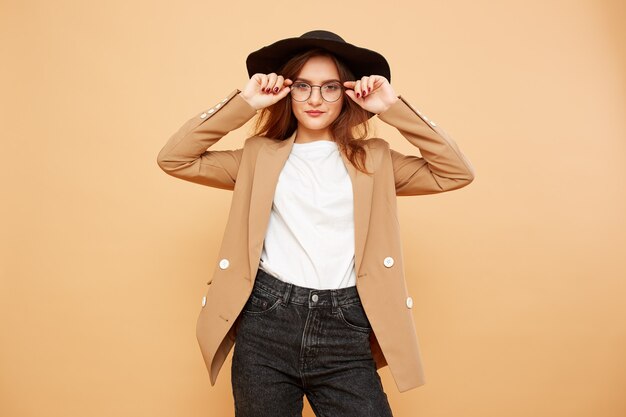 Bella ragazza bruna con i capelli lunghi con gli occhiali e un cappello nero in testa vestita con t-shirt bianca, jeans e giacca beige è in posa sullo sfondo beige in studio.