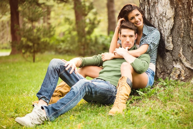 Bella ragazza bruna con un bel ragazzo, seduto sull'erba nel parco. La ragazza era appoggiata a un albero con un sorriso guardando la telecamera, un ragazzo seduto tra le gambe della ragazza, appoggiato all'indietro