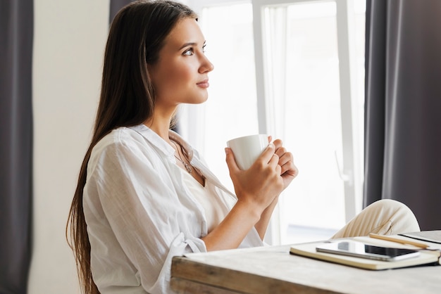 Bella ragazza bruna è seduta al tavolo davanti al computer portatile, tenendo la tazza di caffè con le mani.