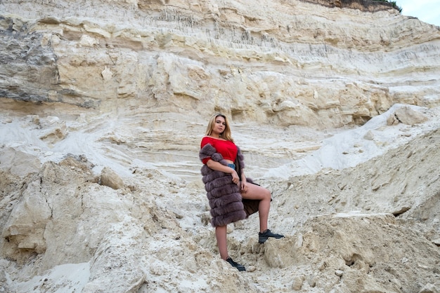 Bella ragazza bruna in maglietta e pantaloncini ocer pelliccia in posa a rocce di sabbia vuote