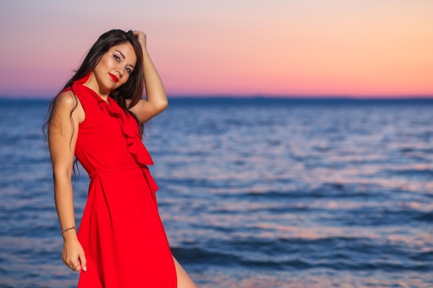 Bella ragazza bruna in un vestito rosso in posa in riva al mare durante l'alba o il tramonto
