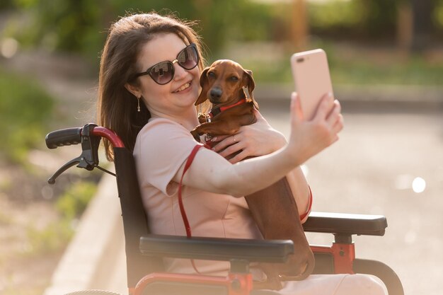 bella ragazza bruna su una sedia a rotelle in estate durante una passeggiata con un simpatico cane bassotto prende una se