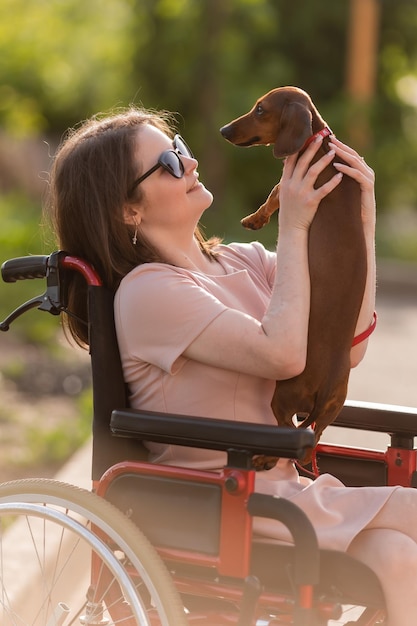 bella ragazza bruna su una sedia a rotelle in estate durante una passeggiata con un simpatico cane bassotto