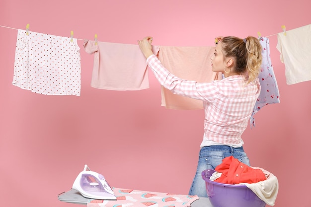 Bella ragazza casalinga in camicia a scacchi vestiti asciutti su corda che stira vestiti puliti mentre fa i lavori domestici isolati su sfondo rosa ritratto in studio. Concetto di pulizia. Mock up spazio di copia.