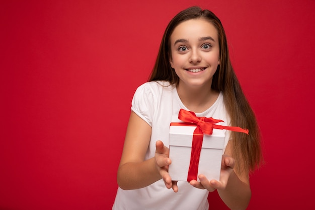 Bella ragazza castana felice isolata sopra la parete variopinta del fondo che porta i vestiti casuali alla moda che tengono il contenitore di regalo