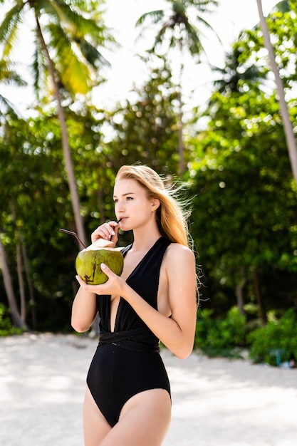 Bella ragazza castana in bikini nero che beve il latte di acqua di cocco vicino all'oceano