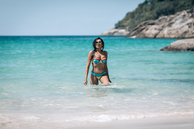 Bella ragazza castana in bikini variopinto e occhiali da sole in piedi in acqua alla spiaggia esotica. Phuket. Tailandia. Bellissimo paesaggio marino sfocato