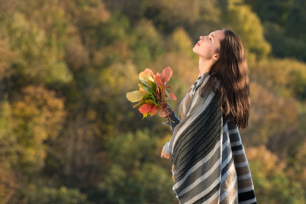 Bella ragazza castana in un poncho che tiene un mazzo di foglie di autunno e guardando il cielo