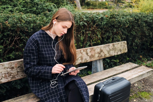 Bella ragazza casual in auricolari utilizzando premurosamente tablet seduto con la valigia sulla panchina nel parco cittadino
