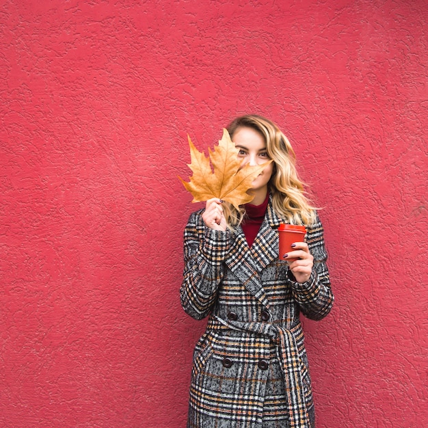 Bella ragazza caucasica che tiene una foglia gialla di autunno e una tazza di caffè rossa