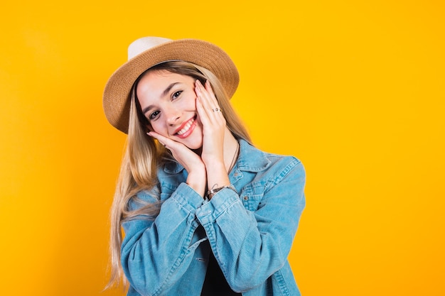 Bella ragazza caucasica con cappello e mani sul viso su sfondo giallo.