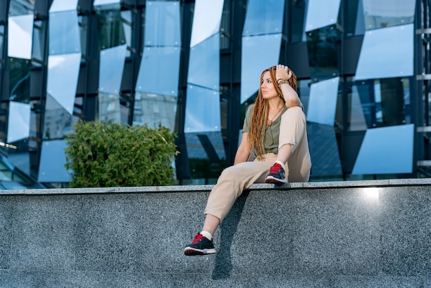 Bella ragazza caucasica con i dreadlocks seduta sul parapetto e guardando in lontananza. Edificio per uffici grigio sullo sfondo