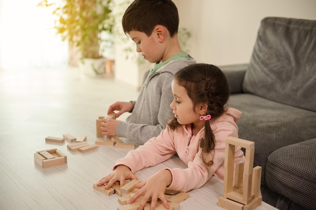 Bella ragazza caucasica curiosa che ispeziona gli edifici in blocchi di legno di suo fratello - attività dell'infanzia, concetto di sviluppo delle abilità motorie. Fratello e sorella che giocano insieme a giochi da tavolo a casa