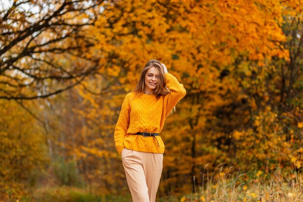 Bella ragazza caucasica felice con un sorriso in un maglione giallo vintage alla moda cammina in un parco autunnale con fogliame dai colori vivaci