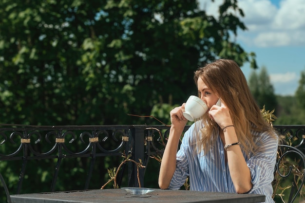 bella ragazza che beve caffè al tavolo sul balcone e parla al telefono