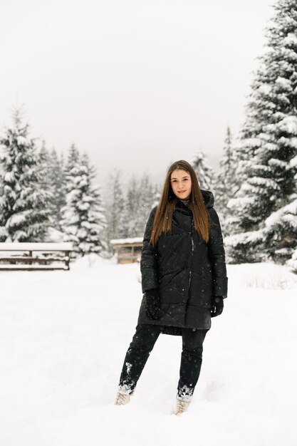 Bella ragazza che cammina in una nevicata. Giovane donna in piumino nero in posa nella foresta d'inverno. Ritratto all'aperto di bella signora bruna. Concetto invernale