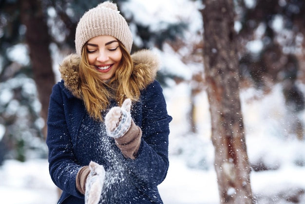 Bella ragazza che gioca con la neve nella foresta d'inverno Ragazza sorridente in una giacca blu e cappello lavorato a maglia