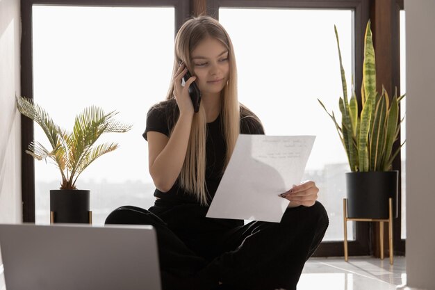 Bella ragazza che lavora a casa al computer seduto sul pavimento vicino alla finestra e parlando al telefono sorridendo