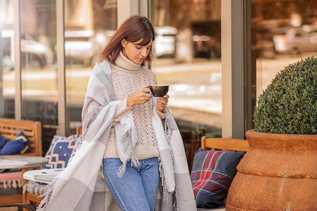 Bella ragazza che riposa e beve caffè seduti al bar sulla terrazza, avvolta in una coperta a quadri di lana. Mattina d'autunno.