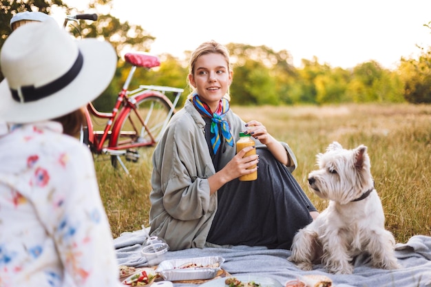 Bella ragazza che tiene in mano una bottiglia di frullato che guarda sognante da parte con un piccolo cane carino vicino al picnic nel parco