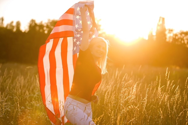 Bella ragazza che tiene una bandiera americana nel vento in un campo di segale. Paesaggio estivo contro il cielo blu. Orientamento orizzontale.